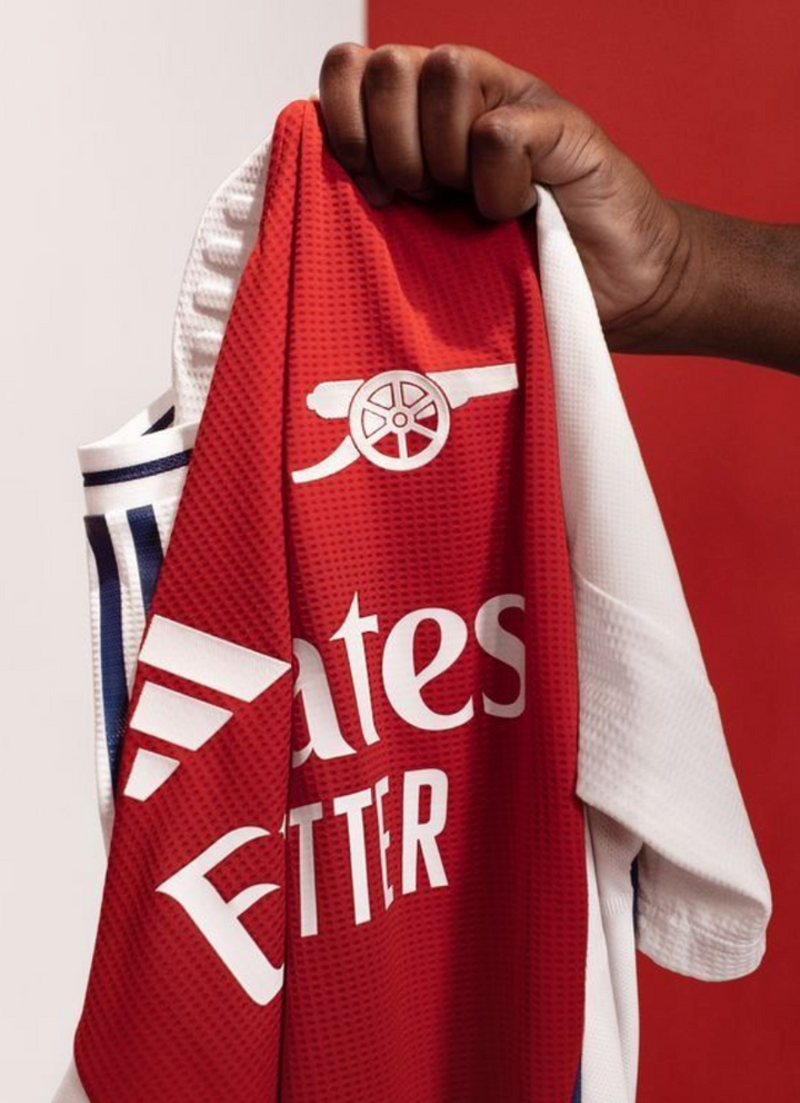 Hand holding the Arsenal 2024 home jersey featuring red and white colors with the club logo
